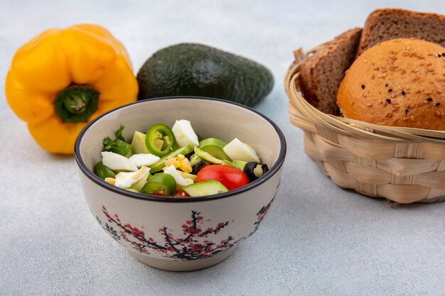 Front view of fresh vegetable salad including pepper cucumber tomato in a bowl with a basket of breads avocado and yellow bell pepper on white surface