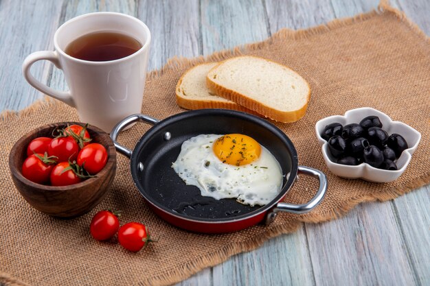 자루와 나무 표면에 빵 조각과 올리브 그릇과 토마토와 차 그릇과 함께 냄비에 튀긴 계란의 전면보기