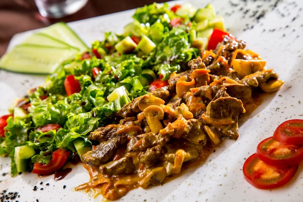 Вид спереди жареного мяса с грибами в соусе с овощным салатом и ломтиками помидора и огурца