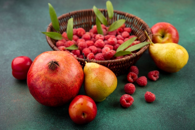 Front view of fruits as pomegranate peach plum with basket of raspberry on green surface