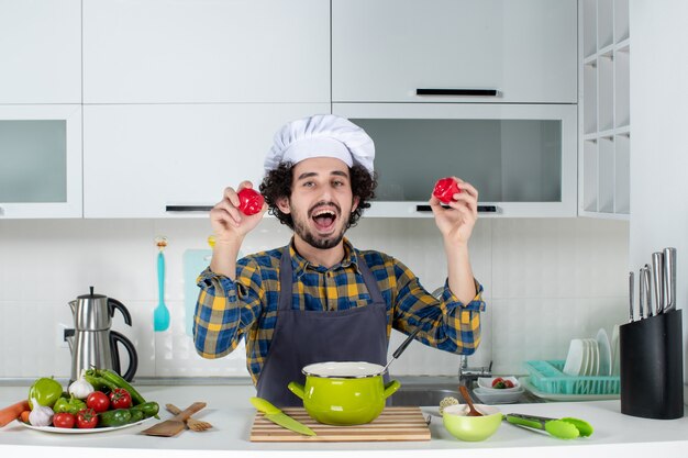 Вид спереди забавного и эмоционального шеф-повара со свежими овощами, держащего красный перец на белой кухне