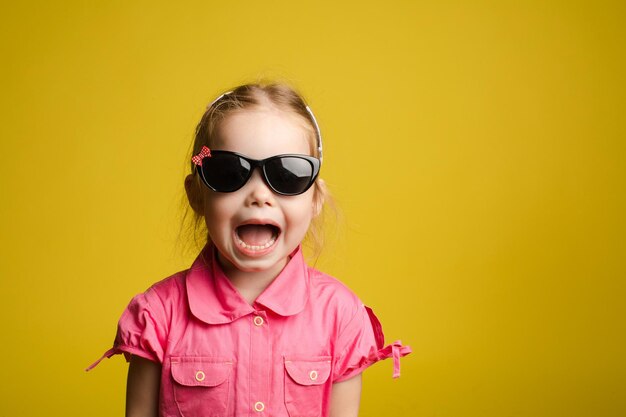 Vista frontale della ragazza divertente in camicia rosa con gli occhiali e gridando grazioso bambino che guarda l'obbiettivo e posa su sfondo giallo isolato concetto di outfit estivo e infanzia