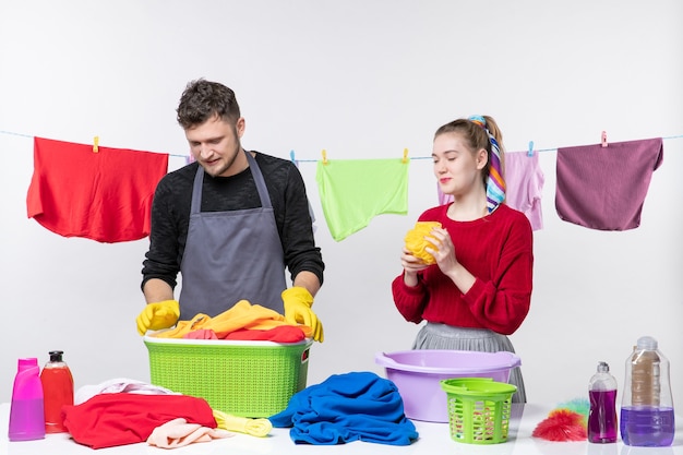 Front view funny young couple man holding laundry basket and wife holding bath sponge on white wall