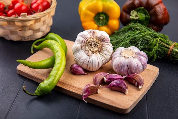 Front view of garlic bulb and cloves pepper on cutting board and basket of tomato pepper bunch of dill on black surface