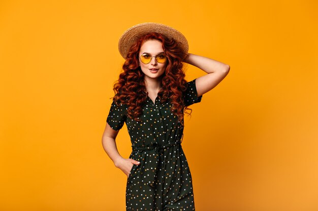 Front view of ginger girl in vintage outfit. Studio shot of pretty young lady in sunglasses and straw hat.