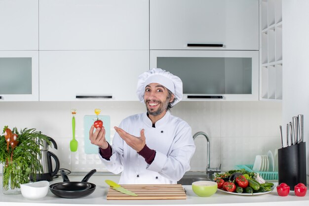 Front view glad male chef in uniform holding up tomato in kitchen