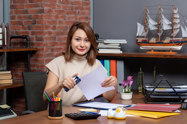 Front view glad woman using stapler sitting at desk in office