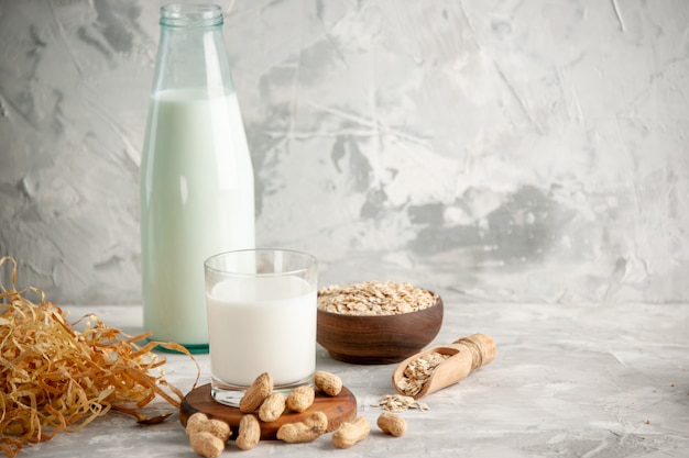 Front view of glass bottle and cup filled with milk on wooden tray and dry fruits spoon oats in brown pot on the left side on white table on ice background