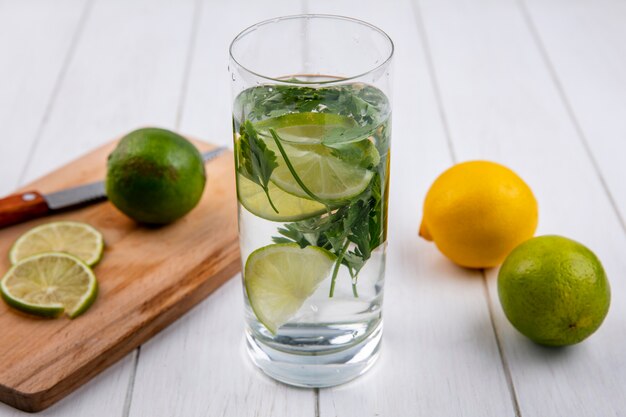 Front view of glass of water with greens and lime and lemon on a blackboard with a knife on a white surface