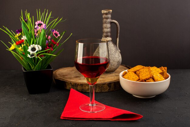 A front view glass of wine along with flower and crisps inside plate on dark desk