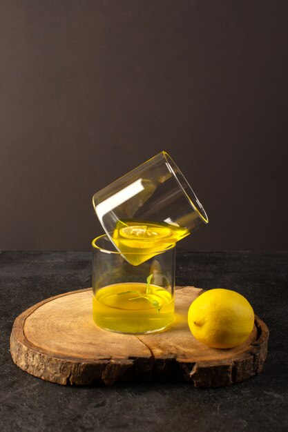 茶色の木製の机の上のレモン全体と灰色の背景のカクテルレモンドリンクに沿って透明なグラスの内側にジュースレモンジュースが入った正面グラス