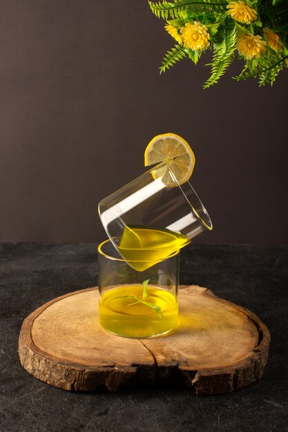 Очки спереди с соком лимонного сока внутри прозрачных стаканов вместе с цветами на коричневом деревянном столе и сером фоне коктейль лимонный напиток