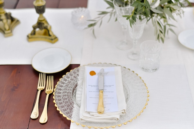 Vista frontale di bicchieri e posate dorate servite sul tavolo di legno e targhetta stampata per gli ospiti e tovaglioli in tessuto bianco