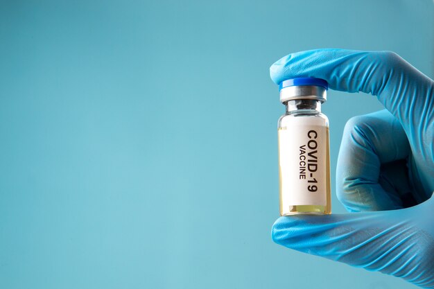 파란색 물결 배경의 왼쪽에 covid-백신이 있는 닫힌 앰플을 들고 있는 장갑의 전면