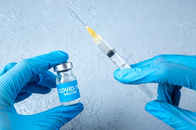 Front view of glove wearing hand holding full syringe and covid- vaccine on gray sand background