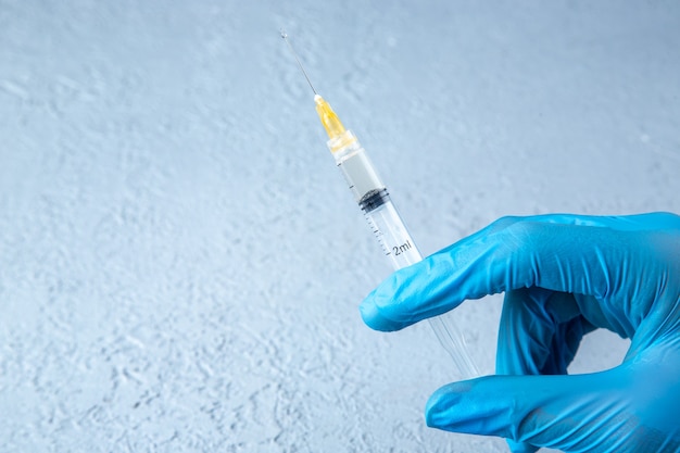 Front view of glove wearing hand holding full syringe on the left side on gray sand background