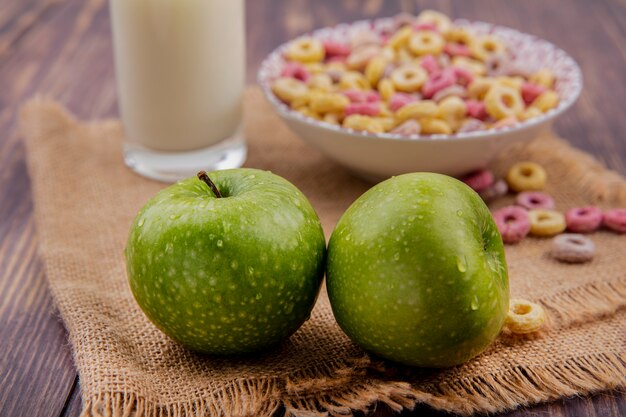 Front view of green apples with cereals on a bowl with a glass of milk on sack cloth on a wooden surface