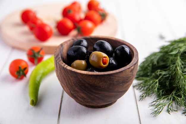 Front view of green and black olives in bowl and tomatoes pepper and dill on wooden surface