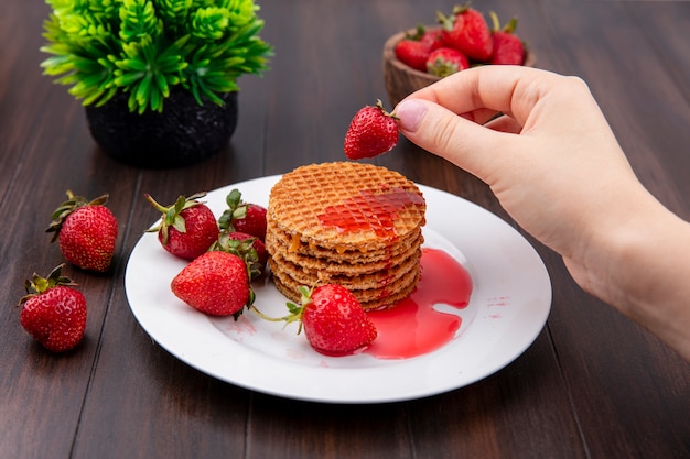 Front view of hand holding strawberry with waffle biscuits in plate and bowl of strawberry and flower on wooden surface