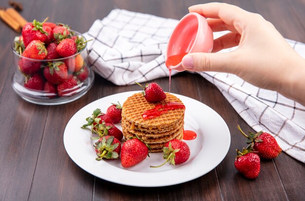 Front view of hand pouring strawberry syrup from bowl onto waffle biscuits and bowl of strawberry on wooden surface