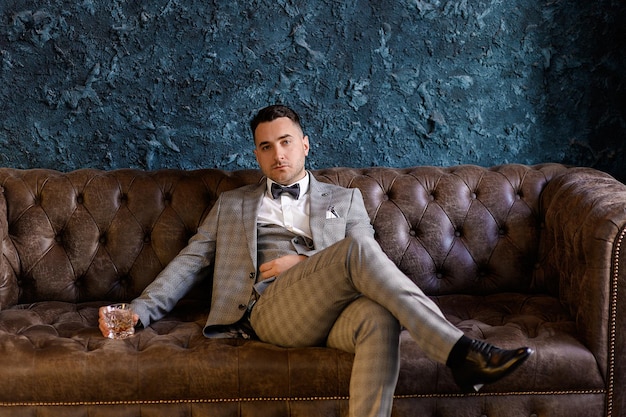 Front view of handsome bridegroom with beard wearing in trendy suit sitting on leather sofa in luxury apartment holding glass filled with beverage and looking at camera