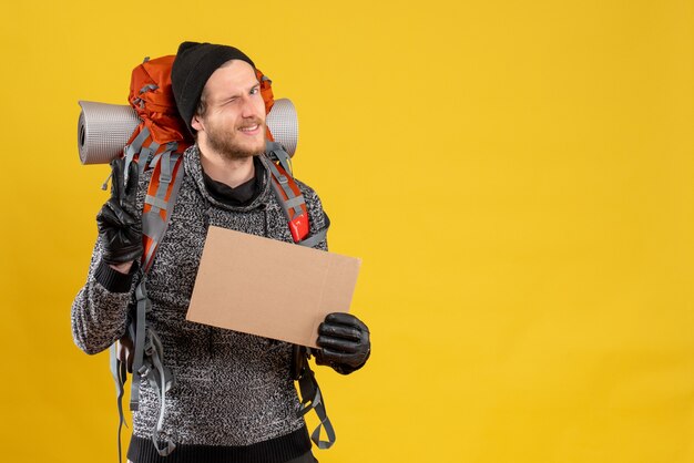 Front view of handsome male hitchhiker with leather gloves and backpack holding blank cardboard gesturing victory sign