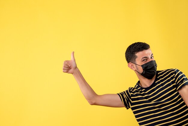 Front view handsome man in black mask making thumb up sign on yellow isolated background