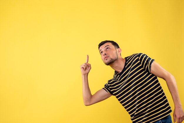 Front view handsome man in black and white striped t-shirt pointing at ceiling on yellow isolated background