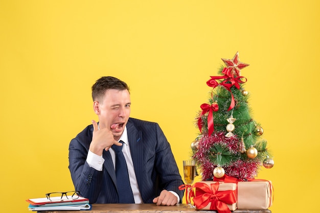 クリスマスツリーと黄色の贈り物の近くのテーブルに座っているカメラで点滅しているハンサムな男の正面図