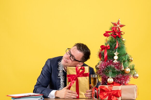Вид спереди красивого мужчины, держащего свой подарок, сидящего за столом возле рождественской елки и подарков на желтом