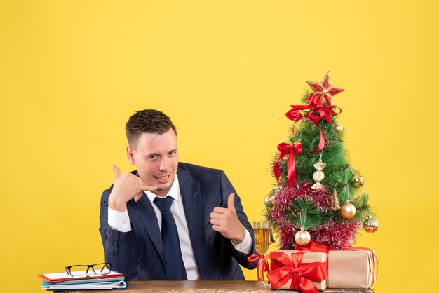 Front view of handsome man making call me phone sign sitting at the table near xmas tree and gifts on yellow