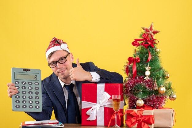 Вид спереди красивого мужчины, делающего большой палец вверх знак, сидящего за столом возле рождественской елки и подарков на желтом