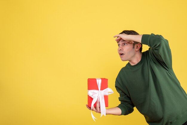 Front view handsome young man holding gift looking at something on yellow 