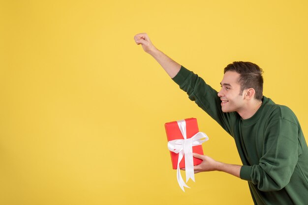 Front view handsome young man holding gift making super hero gesture on yellow 