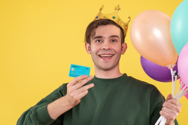 Front view handsome young man with crown holding balloons and card on yellow 