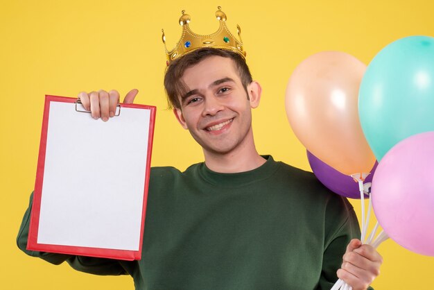 Front view handsome young man with crown holding balloons and clipboard on yellow 