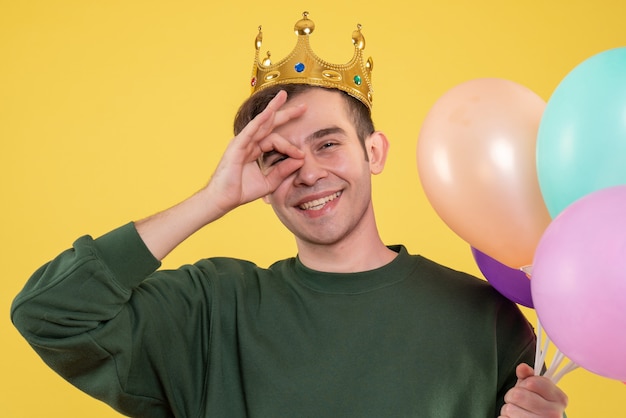 Бесплатное фото Вид спереди красивый молодой человек с короной, держащий воздушные шары, поставив знак ок перед глазом на желтом