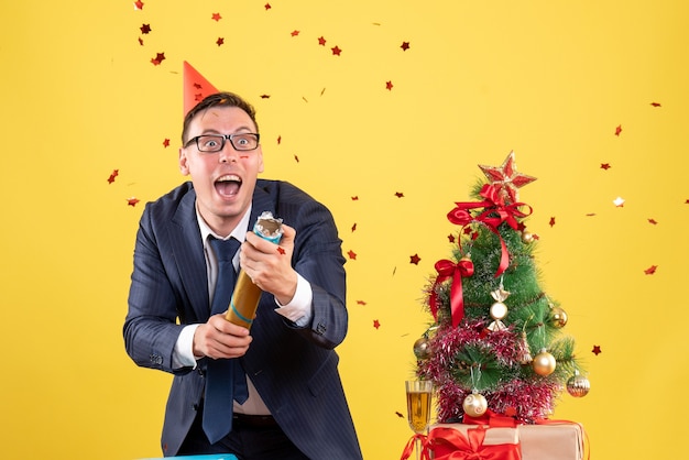 Free photo front view of happy business man using party popper standing behind the table near xmas tree and gifts on yellow