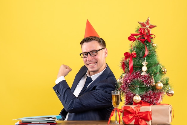 Front view of happy business man with party cap sitting at the table near xmas tree and presents on yellow