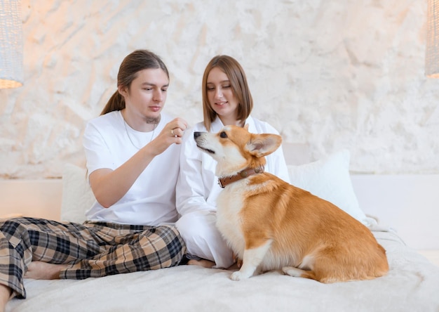 Front view of happy couple resting on bed wearing pajamas and woman admiring and sitting near guy which feeding funny ginger corgi from hand indoors