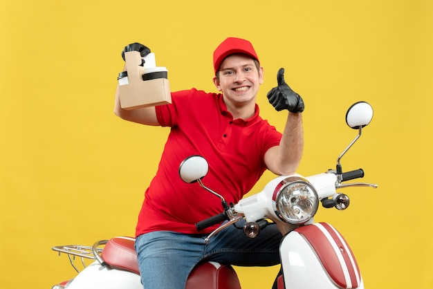 Vista frontale dell'uomo felice del corriere che porta i guanti rossi del cappello e della camicetta nella mascherina medica che trasporta l'ordine che si siede sugli ordini della tenuta dello scooter che indicano verso l'alto