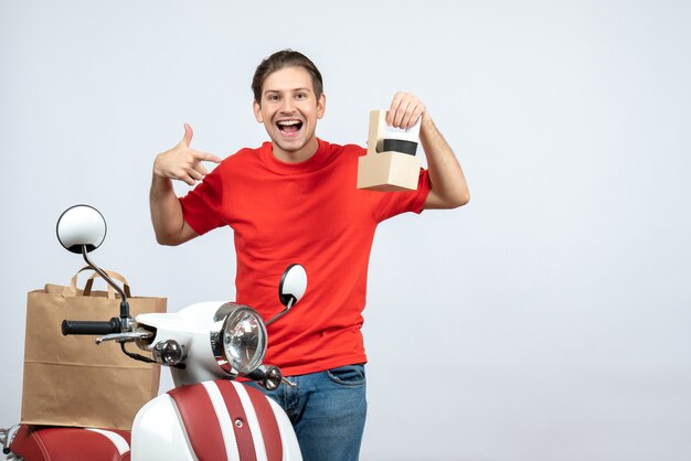 Front view of happy delivery man in red uniform standing near scooter showing order on white background