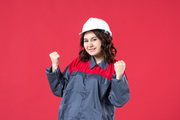 Front view of happy female builder in uniform with hard hat on isolated red background