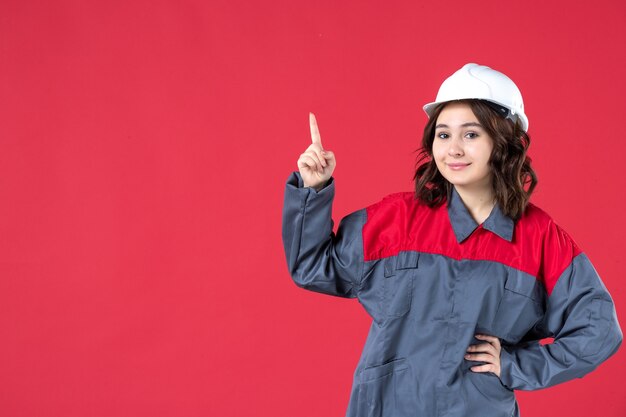 Front view of happy female builder in uniform with hard hat and pointing up on isolated red background