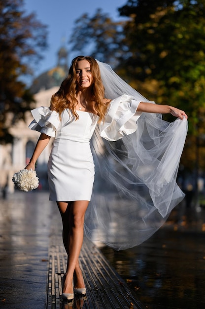 Front view of happy female mode in wedding slim short gown and heels holding bouquet of white flowers and waving long veil while walking in sunny town