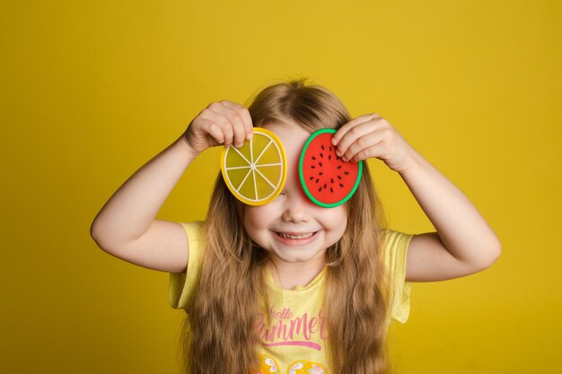 黄色の孤立した背景の上に立っておもちゃで目を閉じる幸せな女の子の正面図笑って遊んでいる陽気な長髪の子供かくれんぼゲームと幸せの概念