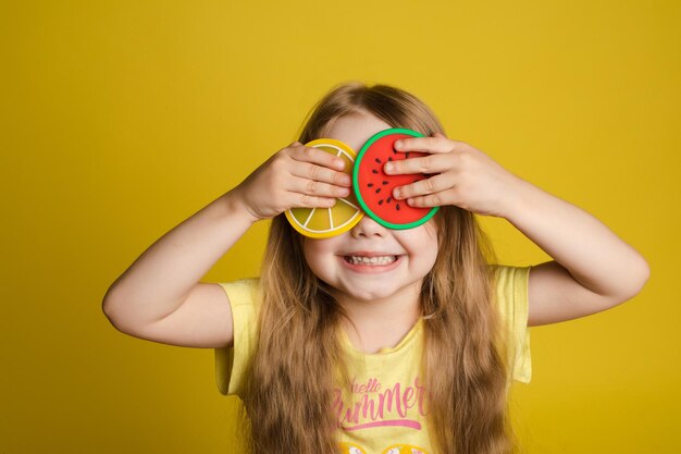 Front view of happy girl standing on yellow isolated background and closing eyes with toys Cheerful longhaired child laughing and playing hide and seek Concept of game and happiness