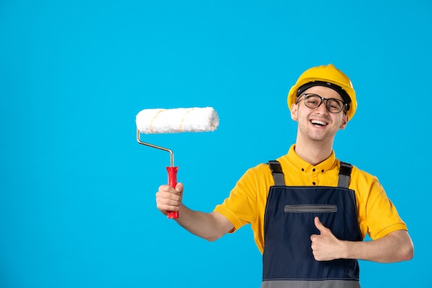 Front view of happy male builder in uniform with paint roller in his hands on blue surface