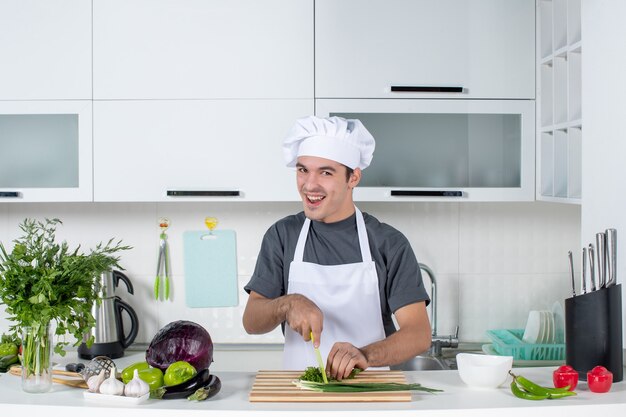 Front view happy male chef in uniform cutting greens on wood board behind kitchen table