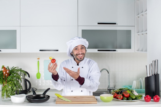 Front view happy male chef in uniform holding up tomato in kitchen